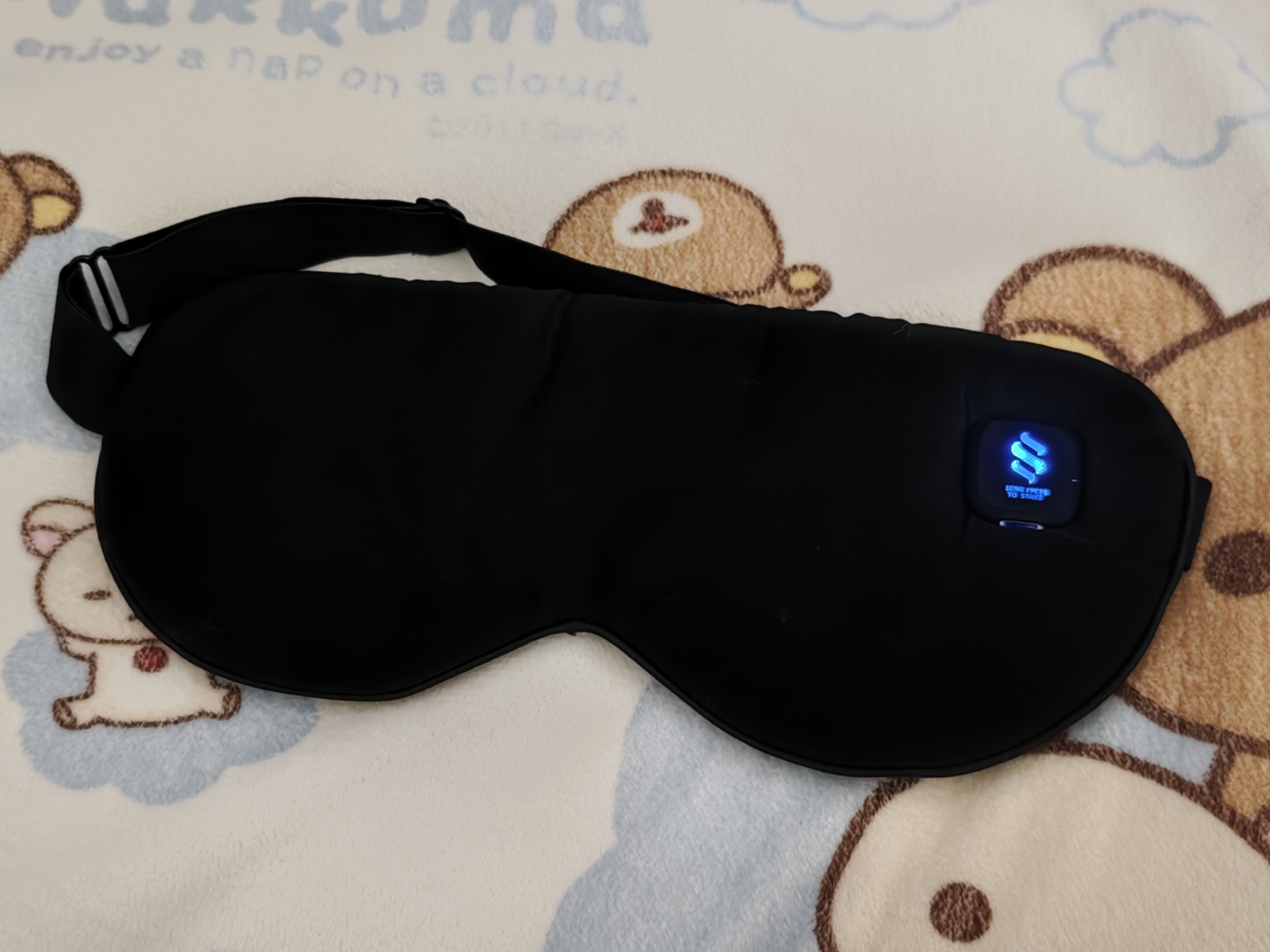 黒いホットアイマスク。 右上に青く光っている（低温の設定）。 背景はリラックマの枕。